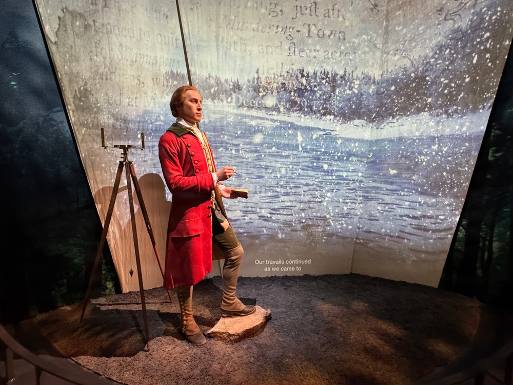 A museum exhibit featuring a wax or animatronic figure dressed in 18th-century colonial attire, standing in a dramatic immersive display with projected imagery of a snowy landscape and historical text. The figure holds a small book or journal and appears to be narrating or recounting a journey. A subtitle on the projection reads 'Our travails continued as we came to,' suggesting this is an interactive or theatrical historical presentation.