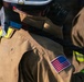 The image shows the back view of one or more firefighters wearing standard turnout gear. The protective clothing is tan/brown in color with yellow reflective striping, and features a prominent American flag patch on the upper right sleeve. A helmet with an attached tool or device is visible at the top of the frame.