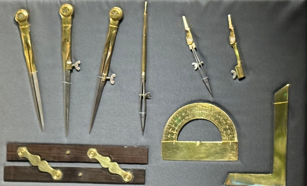 The image shows a collection of antique brass drafting and mathematical instruments displayed on a gray surface. The tools appear to be historical surveying or technical drawing instruments, likely from the 18th or 19th century. Items include various compasses, dividers, a protractor, a set square, and what appear to be parallel rulers or linkage bars.