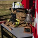 A ceremonial display table featuring firefighter gear and a memorial trophy, likely set up for a fire department ceremony or retirement event. The table holds turnout gear, a white fire helmet, boots, an axe, and a silver bell trophy mounted on a wooden base listing '507th Fire Chiefs'. An American flag is partially visible on the right side of the frame.