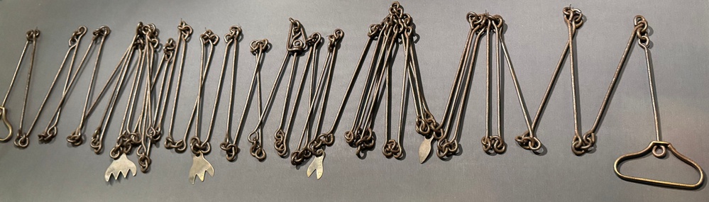 The image shows a collection of metal implements laid out in a row on a gray surface. These appear to be antique or historical metal tools, possibly branding irons, livestock markers, or decorative pins, featuring looped tops and various shaped ends including claw-like or fork-shaped tips. The items are arranged systematically for display or cataloging purposes.