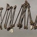 The image shows a collection of metal implements laid out in a row on a gray surface. These appear to be antique or historical metal tools, possibly branding irons, livestock markers, or decorative pins, featuring looped tops and various shaped ends including claw-like or fork-shaped tips. The items are arranged systematically for display or cataloging purposes.