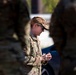 A female military service member in OCP (Operational Camouflage Pattern) uniform is photographed in profile, appearing to look down at her hands. She is framed between two out-of-focus figures in matching camouflage uniforms in the foreground. The setting appears to be an outdoor military environment with vehicles visible in the background.