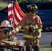 A firefighter in full turnout gear, wearing a black helmet labeled 'Firefighter,' stands beside a table displaying a set of folded firefighting gear topped with a white helmet. An American flag is prominently displayed in the background, suggesting a memorial or ceremonial event. The scene appears to be an outdoor tribute, possibly honoring a fallen firefighter.