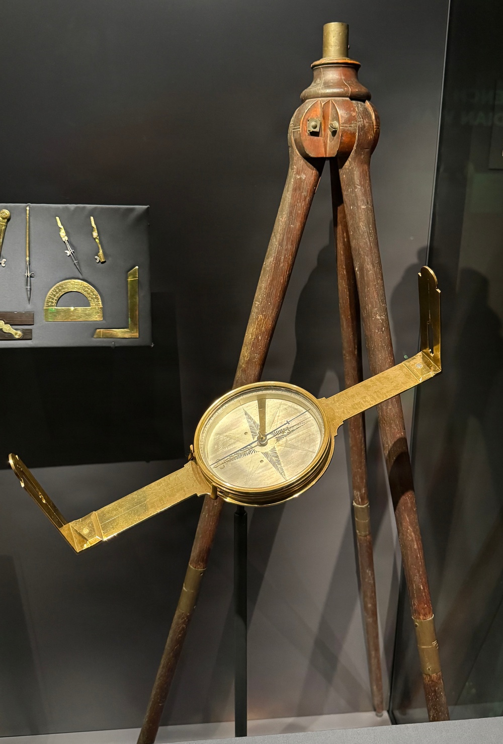 A museum display featuring an antique surveying compass (circumferentor) mounted on a wooden tripod stand. The brass instrument has a circular compass housing with sighting vanes extending horizontally on both sides. In the background, a secondary display case shows smaller brass drafting and mathematical instruments.