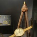 A museum display featuring an antique surveying compass (circumferentor) mounted on a wooden tripod stand. The brass instrument has a circular compass housing with sighting vanes extending horizontally on both sides. In the background, a secondary display case shows smaller brass drafting and mathematical instruments.