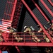A group of firefighters in full turnout gear and SCBA (self-contained breathing apparatus) tanks are climbing a red metal training tower structure. An American flag is prominently displayed on the left side of the structure. The scene appears to depict a firefighter training exercise or a memorial stair climb event.