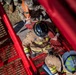 An aerial/overhead view of firefighters in full turnout gear working alongside military personnel in camouflage uniforms. The firefighters appear to be handling coiled fire hoses on what appears to be a red fire truck or apparatus. The scene suggests a joint training exercise between civilian firefighters and military units.