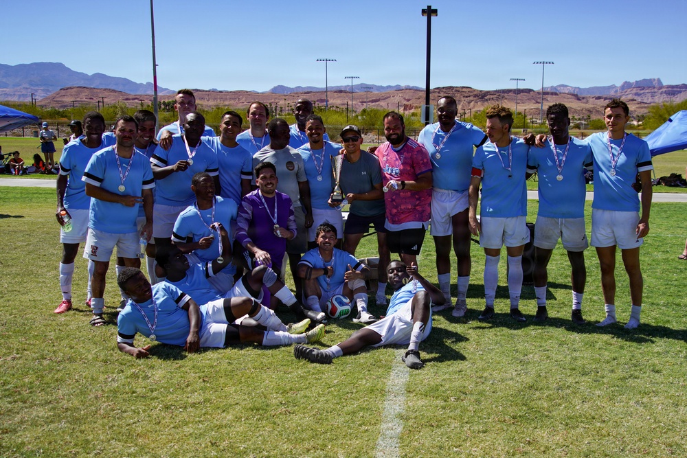 A soccer team poses for a group photo on a grass field after winning a tournament, with most players wearing light blue and white uniforms and medals around their necks. One person in the center is holding a trophy cup, and the team is arranged in three rows — standing, kneeling, and lying on the ground. The setting appears to be an outdoor sports complex in a desert region, with red rock mountains visible in the background.