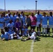 A soccer team poses for a group photo on a grass field after winning a tournament, with most players wearing light blue and white uniforms and medals around their necks. One person in the center is holding a trophy cup, and the team is arranged in three rows — standing, kneeling, and lying on the ground. The setting appears to be an outdoor sports complex in a desert region, with red rock mountains visible in the background.