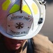 A close-up photograph of a white firefighter's helmet belonging to an Assistant Chief at Tinker Air Force Base (AFB). The helmet features a shield-shaped badge with crossed fire axes/tools and high-visibility yellow-green reflective tape strips on top. The wearer, dressed in tan turnout gear, is looking downward.
