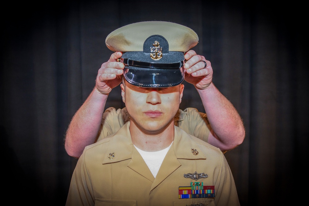 A U.S. Navy Chief Petty Officer is having a combination cover (white-topped service cap) placed on his head during what appears to be a Chief Petty Officer pinning or advancement ceremony. The subject is wearing the tan/khaki Navy Service Uniform with visible rank insignia, warfare device, and service ribbons. Another person's hands are visible placing the cover on his head from behind.