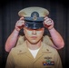 A U.S. Navy Chief Petty Officer is having a combination cover (white-topped service cap) placed on his head during what appears to be a Chief Petty Officer pinning or advancement ceremony. The subject is wearing the tan/khaki Navy Service Uniform with visible rank insignia, warfare device, and service ribbons. Another person's hands are visible placing the cover on his head from behind.
