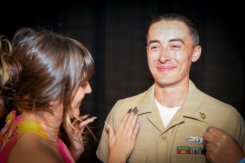 A U.S. Navy servicemember in khaki uniform is visibly emotional, appearing to have tears in his eyes, while a woman pins or adjusts a badge/insignia on his chest. The scene appears to be a military pinning ceremony, a significant milestone event. The woman has decorative patriotic-themed nail art and is wearing a colorful dress.