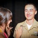 A U.S. Navy servicemember in khaki uniform is visibly emotional, appearing to have tears in his eyes, while a woman pins or adjusts a badge/insignia on his chest. The scene appears to be a military pinning ceremony, a significant milestone event. The woman has decorative patriotic-themed nail art and is wearing a colorful dress.