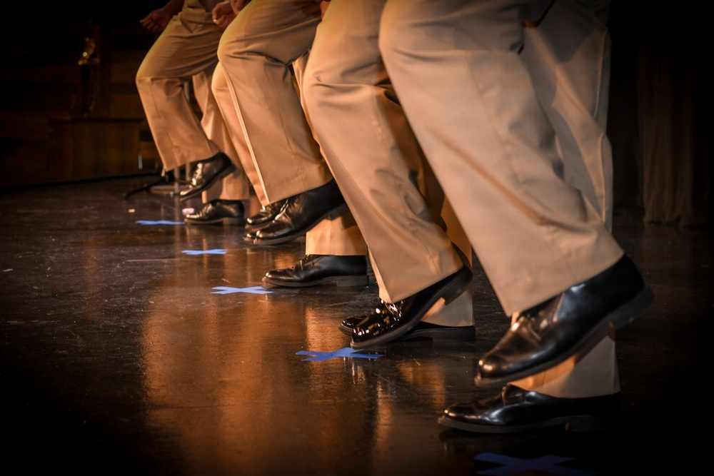 The image shows a close-up view of multiple performers' legs and feet on a stage, all wearing matching tan/khaki trousers and polished black dress shoes. The individuals appear to be in a synchronized pose, with knees slightly bent and feet positioned similarly, suggesting a choreographed performance. Blue tape marks are visible on the dark wooden stage floor, likely serving as position markers.