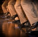The image shows a close-up view of multiple performers' legs and feet on a stage, all wearing matching tan/khaki trousers and polished black dress shoes. The individuals appear to be in a synchronized pose, with knees slightly bent and feet positioned similarly, suggesting a choreographed performance. Blue tape marks are visible on the dark wooden stage floor, likely serving as position markers.