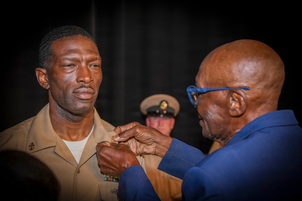 A military promotion or award ceremony is taking place, where an older civilian man in a blue suit is pinning an insignia or device onto the collar/chest area of a U.S. Navy enlisted service member in tan/khaki uniform. The service member stands at attention with a solemn expression. A third individual in a military cover (cap) is visible in the background.