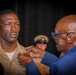 A military promotion or award ceremony is taking place, where an older civilian man in a blue suit is pinning an insignia or device onto the collar/chest area of a U.S. Navy enlisted service member in tan/khaki uniform. The service member stands at attention with a solemn expression. A third individual in a military cover (cap) is visible in the background.