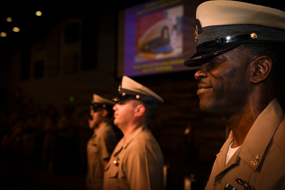 Three military personnel in U.S. Navy dress uniforms stand at attention in a formal indoor setting, likely during a ceremony or official event. They are wearing khaki service uniforms with combination covers (peaked caps) bearing Navy insignia. A projection screen displaying what appears to be a Navy-related image is visible in the background.