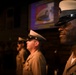 Three military personnel in U.S. Navy dress uniforms stand at attention in a formal indoor setting, likely during a ceremony or official event. They are wearing khaki service uniforms with combination covers (peaked caps) bearing Navy insignia. A projection screen displaying what appears to be a Navy-related image is visible in the background.