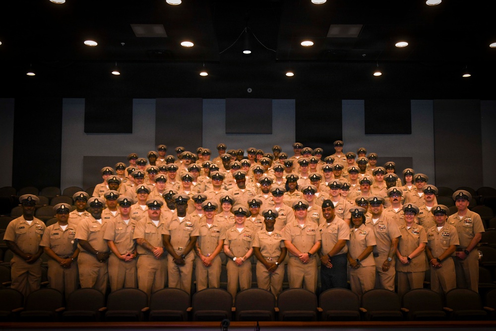 A large group of U.S. Navy personnel in khaki service uniforms are posed for a formal group photograph inside what appears to be an auditorium or theater. The personnel are arranged in multiple tiered rows, with those in the back rows standing on elevated seating levels. All individuals are wearing matching khaki uniforms with combination covers (peaked caps), indicating a formal military gathering or class photo.