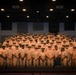 A large group of U.S. Navy personnel in khaki service uniforms are posed for a formal group photograph inside what appears to be an auditorium or theater. The personnel are arranged in multiple tiered rows, with those in the back rows standing on elevated seating levels. All individuals are wearing matching khaki uniforms with combination covers (peaked caps), indicating a formal military gathering or class photo.