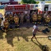 A firefighting training session is taking place outdoors near a red United States Air Force fire truck. An instructor in civilian clothes addresses a large group consisting of personnel in full firefighting turnout gear and military personnel in camouflage uniforms. Breathing apparatus (SCBA) equipment is laid out on the ground in the foreground.