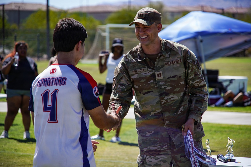 A U.S. Air Force officer identified as 'DeLucia' by his name tape is shaking hands and smiling with a youth athlete wearing a 'Spartans #4' jersey at an outdoor sports event. The setting appears to be a military base sports field where an awards ceremony is taking place, with trophies and medals visible on a table nearby. The interaction suggests the officer is presenting awards or congratulating participants in a youth sports program.
