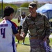 A U.S. Air Force officer identified as 'DeLucia' by his name tape is shaking hands and smiling with a youth athlete wearing a 'Spartans #4' jersey at an outdoor sports event. The setting appears to be a military base sports field where an awards ceremony is taking place, with trophies and medals visible on a table nearby. The interaction suggests the officer is presenting awards or congratulating participants in a youth sports program.