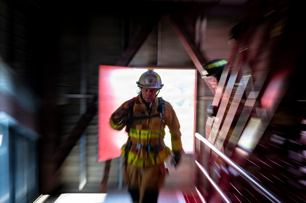 A firefighter in full turnout gear and white helmet is ascending a staircase, framed by a bright red-lit doorway or opening behind them. The image is captured with a zoom burst or radial blur photographic technique, creating a dramatic sense of motion and urgency. The structural elements of what appears to be a training facility or building stairwell are visible around the subject.