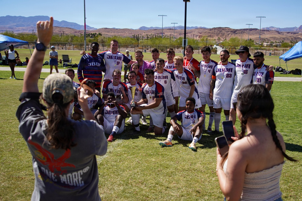 A soccer team wearing white uniforms with 'JBSA' lettering poses for a group photo on a grass field, apparently celebrating a victory as one player holds a trophy. Two people in the foreground are photographing the team, with one raising a hand in celebration. The setting appears to be an outdoor sports complex in a desert region, with red rock mountains visible in the background.