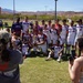 A soccer team wearing white uniforms with 'JBSA' lettering poses for a group photo on a grass field, apparently celebrating a victory as one player holds a trophy. Two people in the foreground are photographing the team, with one raising a hand in celebration. The setting appears to be an outdoor sports complex in a desert region, with red rock mountains visible in the background.