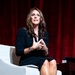 A woman is seated in a white upholstered chair on what appears to be a stage or formal presentation setting, engaged in conversation or a speaking engagement. She is dressed in a dark navy/black outfit and is gesturing with her hands, suggesting active dialogue. The backdrop consists of deep red curtains, and a small table with objects is visible to her right.