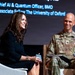 Two individuals are seated on a stage during what appears to be a panel discussion or conference. A woman in a black dress is speaking, while a U.S. Army officer in camouflage uniform listens and smiles. A presentation screen in the background identifies one speaker as 'Chief AI &amp; Quantum Officer, BMO' and 'Associate Fellow, The University of Oxford.'