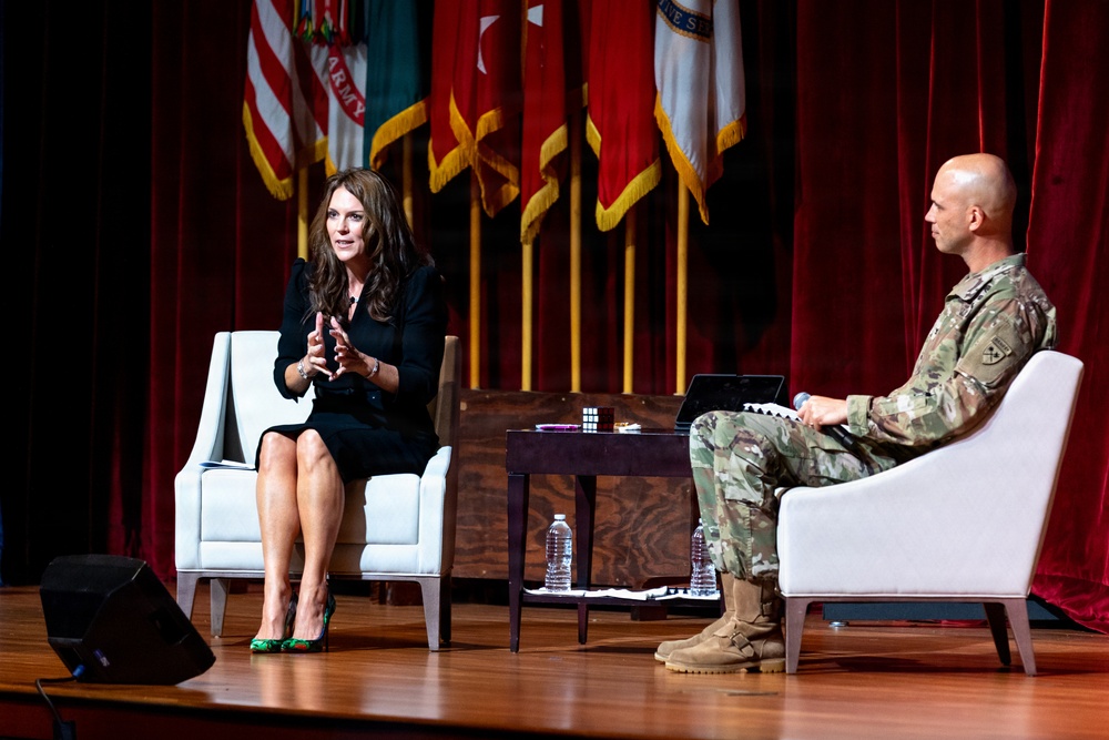 Two individuals are seated on a stage in a panel-style discussion format. A woman in a black outfit is speaking animatedly while a male military officer in camouflage uniform listens. The setting appears to be a formal military event or conference, with multiple military flags displayed in the background.