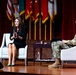 Two individuals are seated on a stage in a panel-style discussion format. A woman in a black outfit is speaking animatedly while a male military officer in camouflage uniform listens. The setting appears to be a formal military event or conference, with multiple military flags displayed in the background.