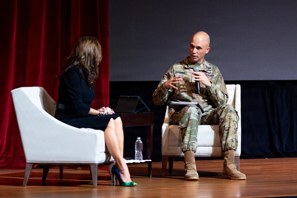 A U.S. Army soldier identified by name tape as 'Carroll' is seated on stage holding a microphone and speaking during what appears to be a formal panel discussion or interview event. He is in conversation with a woman dressed in a black outfit who is seated across from him. The setting appears to be an auditorium or theater with a red curtain backdrop and wooden stage floor.
