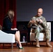 A U.S. Army soldier identified by name tape as 'Carroll' is seated on stage holding a microphone and speaking during what appears to be a formal panel discussion or interview event. He is in conversation with a woman dressed in a black outfit who is seated across from him. The setting appears to be an auditorium or theater with a red curtain backdrop and wooden stage floor.