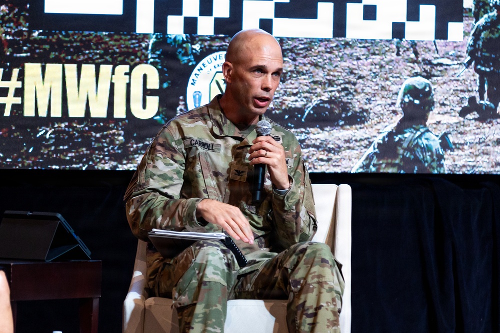 A U.S. Army Colonel named Carroll, wearing Operational Camouflage Pattern (OCP) uniform, is seated in a white chair and speaking into a microphone at what appears to be the Maneuver Warfighter Conference (#MWfC). He is holding papers/notes in his lap and appears to be participating in a panel discussion or presentation. The event backdrop displays the conference hashtag and imagery of soldiers in the field.