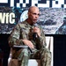 A U.S. Army Colonel named Carroll, wearing Operational Camouflage Pattern (OCP) uniform, is seated in a white chair and speaking into a microphone at what appears to be the Maneuver Warfighter Conference (#MWfC). He is holding papers/notes in his lap and appears to be participating in a panel discussion or presentation. The event backdrop displays the conference hashtag and imagery of soldiers in the field.