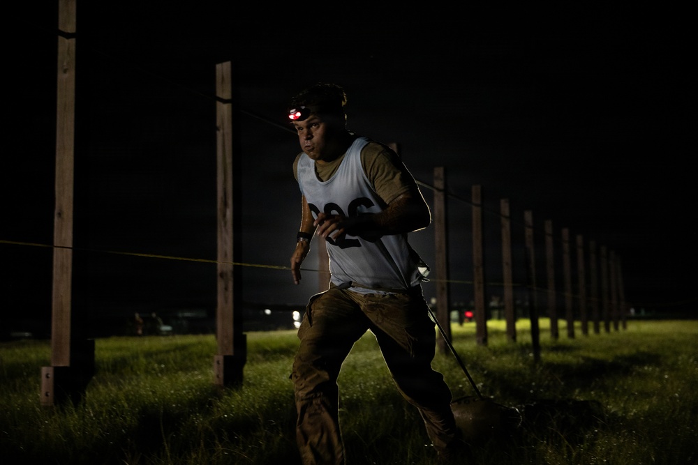 A person is navigating a nighttime obstacle course or endurance race, wearing a numbered bib (appearing to show '226') and a red-lit headlamp. They are moving through a field lined with wooden posts connected by ropes or wires, with mud visible on their clothing. The dramatic lighting and determined expression suggest an intense competitive or training event.