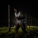 A person is navigating a nighttime obstacle course or endurance race, wearing a numbered bib (appearing to show '226') and a red-lit headlamp. They are moving through a field lined with wooden posts connected by ropes or wires, with mud visible on their clothing. The dramatic lighting and determined expression suggest an intense competitive or training event.