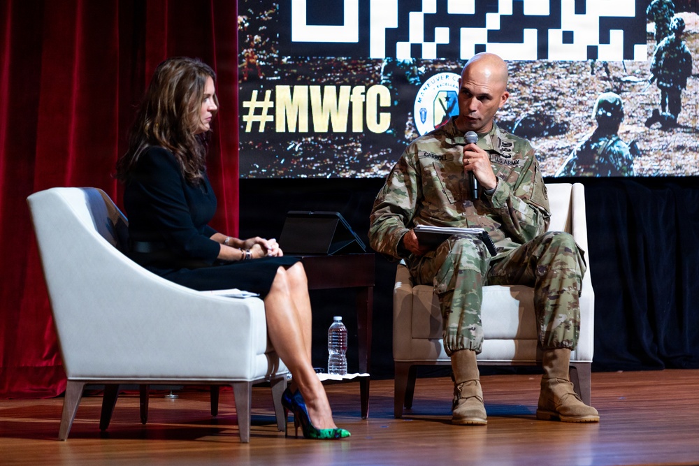 A U.S. Army officer in camouflage uniform is speaking into a microphone while seated on stage alongside a woman in a black dress during what appears to be a panel discussion or interview. The backdrop displays the hashtag '#MWfC' along with military imagery and logos, suggesting a military-themed conference or forum. Both individuals are seated in white armchairs on a wooden stage.