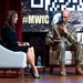 A U.S. Army officer in camouflage uniform is speaking into a microphone while seated on stage alongside a woman in a black dress during what appears to be a panel discussion or interview. The backdrop displays the hashtag '#MWfC' along with military imagery and logos, suggesting a military-themed conference or forum. Both individuals are seated in white armchairs on a wooden stage.