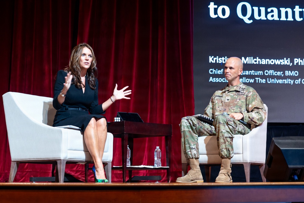 Two individuals are seated on a stage during what appears to be a panel discussion or conference presentation. A woman in a black dress is speaking animatedly while a U.S. Army soldier in camouflage uniform listens. The backdrop screen partially displays text referencing 'Quantum' and identifies 'Kristin Milchanowski, Ph[D]' as 'Chief Quantum Officer, BMO' and 'Associate Fellow, The University of C[hicago?]'.