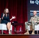 Two individuals are seated on a stage during what appears to be a panel discussion or conference presentation. A woman in a black dress is speaking animatedly while a U.S. Army soldier in camouflage uniform listens. The backdrop screen partially displays text referencing 'Quantum' and identifies 'Kristin Milchanowski, Ph[D]' as 'Chief Quantum Officer, BMO' and 'Associate Fellow, The University of C[hicago?]'.