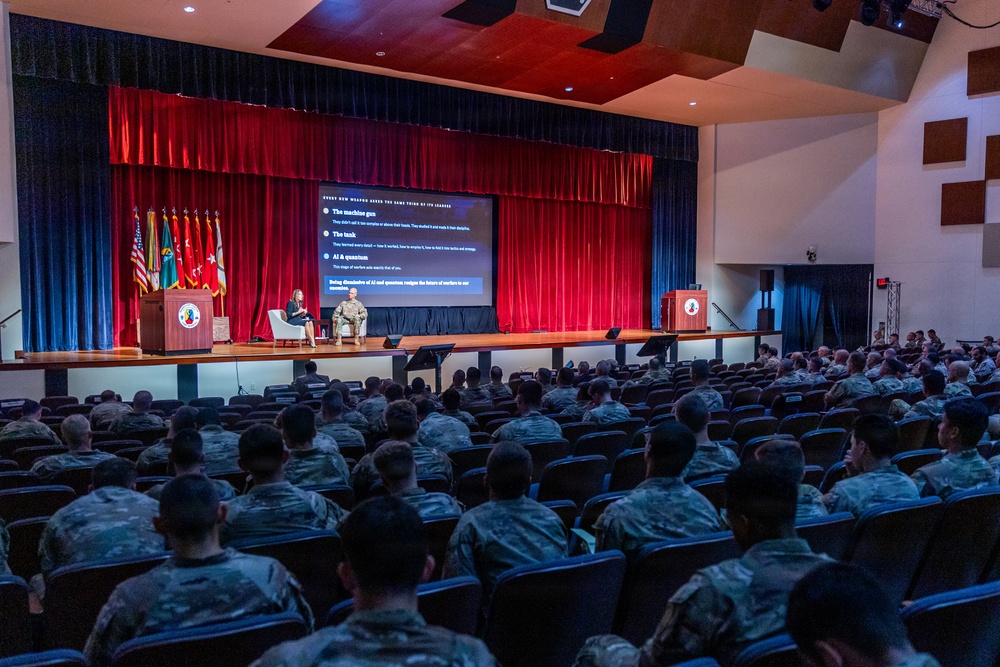 A military briefing or lecture is taking place in a large auditorium filled with uniformed soldiers in camouflage fatigues. On stage, two individuals — one in civilian attire and one in military uniform — are seated in chairs conducting what appears to be a panel discussion or interview-style presentation. A large projection screen behind them displays a slide with bullet points referencing 'The machine gun,' 'The tank,' and 'AI &amp; quantum.'