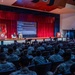 A military briefing or lecture is taking place in a large auditorium filled with uniformed soldiers in camouflage fatigues. On stage, two individuals — one in civilian attire and one in military uniform — are seated in chairs conducting what appears to be a panel discussion or interview-style presentation. A large projection screen behind them displays a slide with bullet points referencing 'The machine gun,' 'The tank,' and 'AI &amp; quantum.'