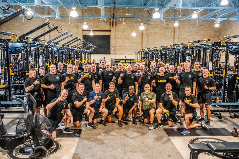 A large group of approximately 40-50 individuals, predominantly wearing black military-style athletic shirts with 'ARMY' branding, pose for a group photo inside a well-equipped military fitness facility. Many individuals are making 'number one' finger gestures toward the camera, suggesting a celebratory or team-unity moment. The facility features rows of black and yellow Rogue fitness equipment including power racks, cable machines, and assault bikes.
